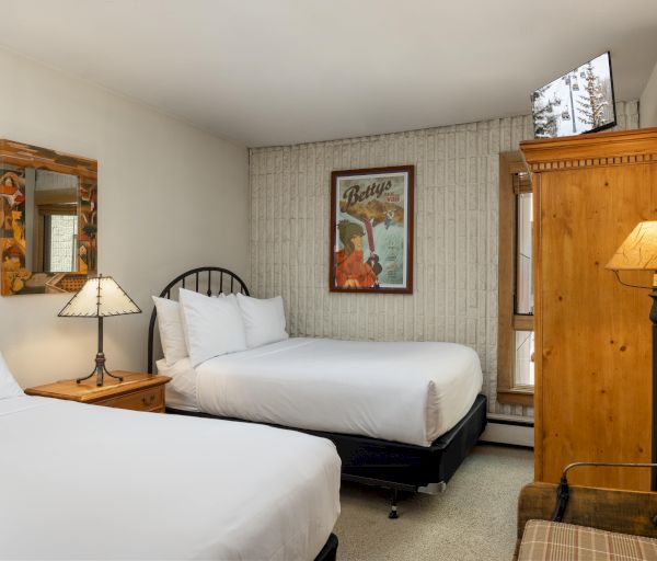 The image shows a cozy hotel room with two beds, a wooden wardrobe, lamps, and a framed wall artwork.