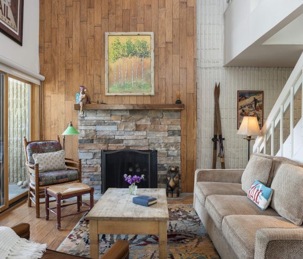 A cozy living room with a stone fireplace, wooden walls, and a sliding door opening to a patio. Decor includes a rug and framed art.