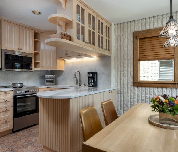 A cozy kitchen with wooden cabinets, stainless steel appliances, a dining table with chairs, and a fruit centerpiece near a window.