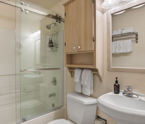 A small bathroom with a glass shower door, sink, mirror, cabinet, and toiletries on a counter, with towels on a rack and toilet.