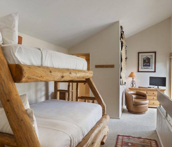 The image shows a cozy room with wooden bunk beds, a small desk, chair, lamp, TV, and large window, creating a welcoming atmosphere.