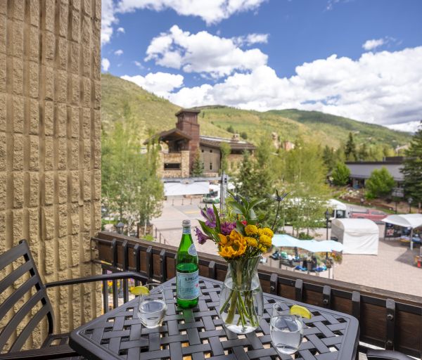 A patio with a table, flowers, and drinks overlooks a scenic view with buildings, trees, and mountains under a cloudy sky.