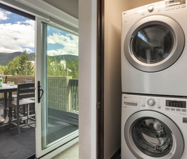 A stacked washer and dryer are next to a sliding glass door leading to a balcony with outdoor seating and a mountain view.