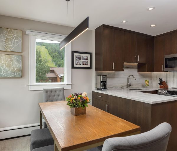 A modern kitchen with dark cabinets, a wooden dining table, and a window view of greenery, featuring minimalist decor and artwork.