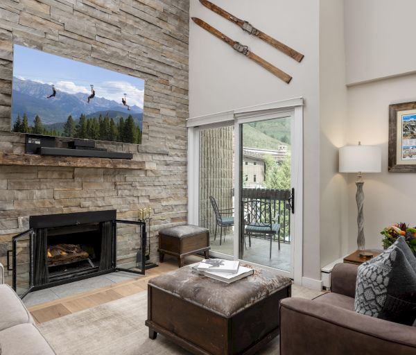 A cozy living room features a stone fireplace, TV, rustic decor with skis, and a view through glass doors leading to a balcony.