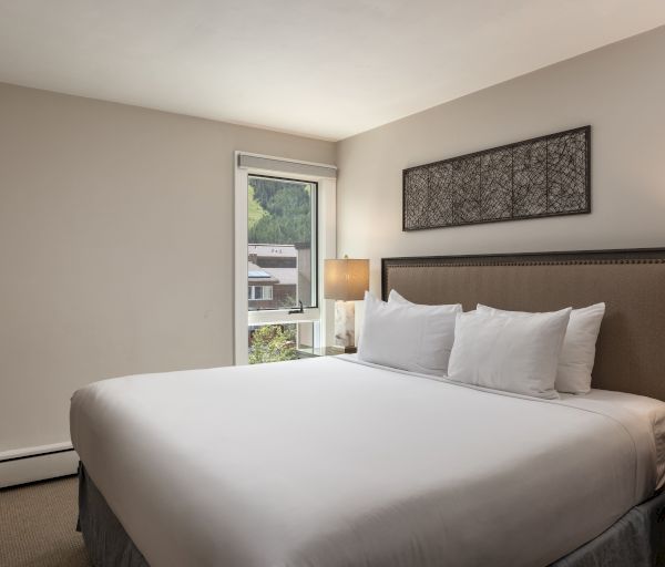 A neatly made bed with white linens is in a hotel room featuring a headboard, wall art, lamps, and a window letting in natural light.