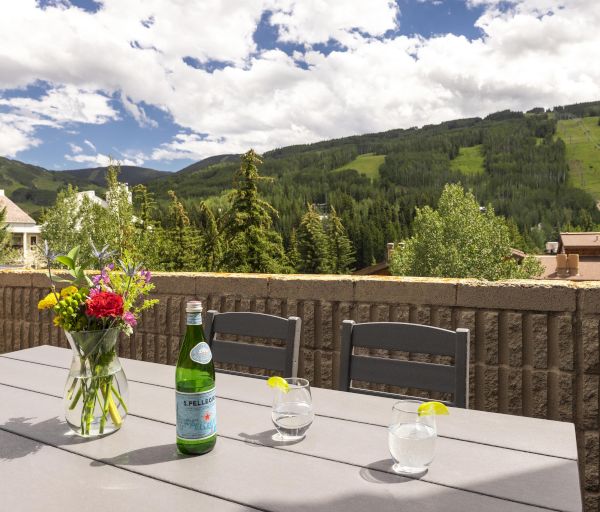 A scenic patio with a table, flowers, a bottle, and glasses overlooks lush green hills and a partly cloudy sky.