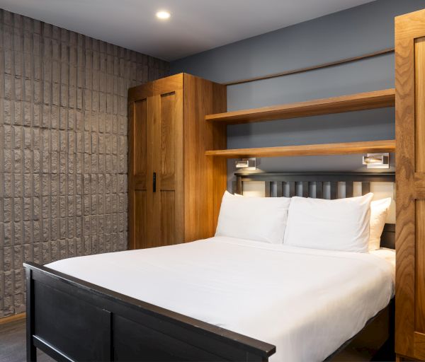 The image shows a modern bedroom with a bed, wooden headboard, shelves, built-in lights, and brick-textured walls, creating a cozy ambiance.