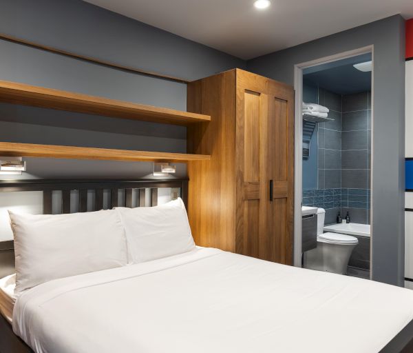 The image shows a modern hotel room featuring a bed, wooden shelves, and a small bathroom area enclosed by a colorful wall design.