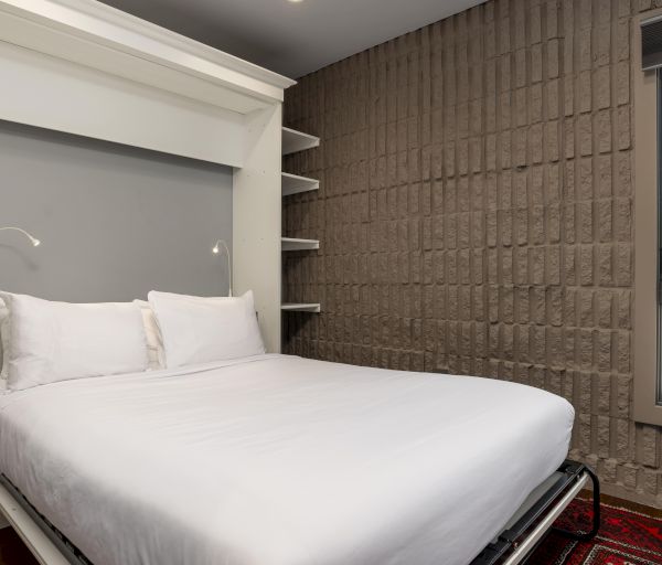 The image shows a room with a wall bed, shelves, gray wall, window with a view, and a red-patterned rug on the floor.