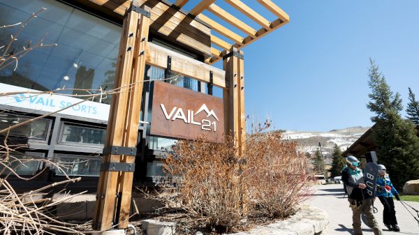 The image shows a building with a sign reading "Vail 21" and people carrying ski equipment outside under a clear blue sky.