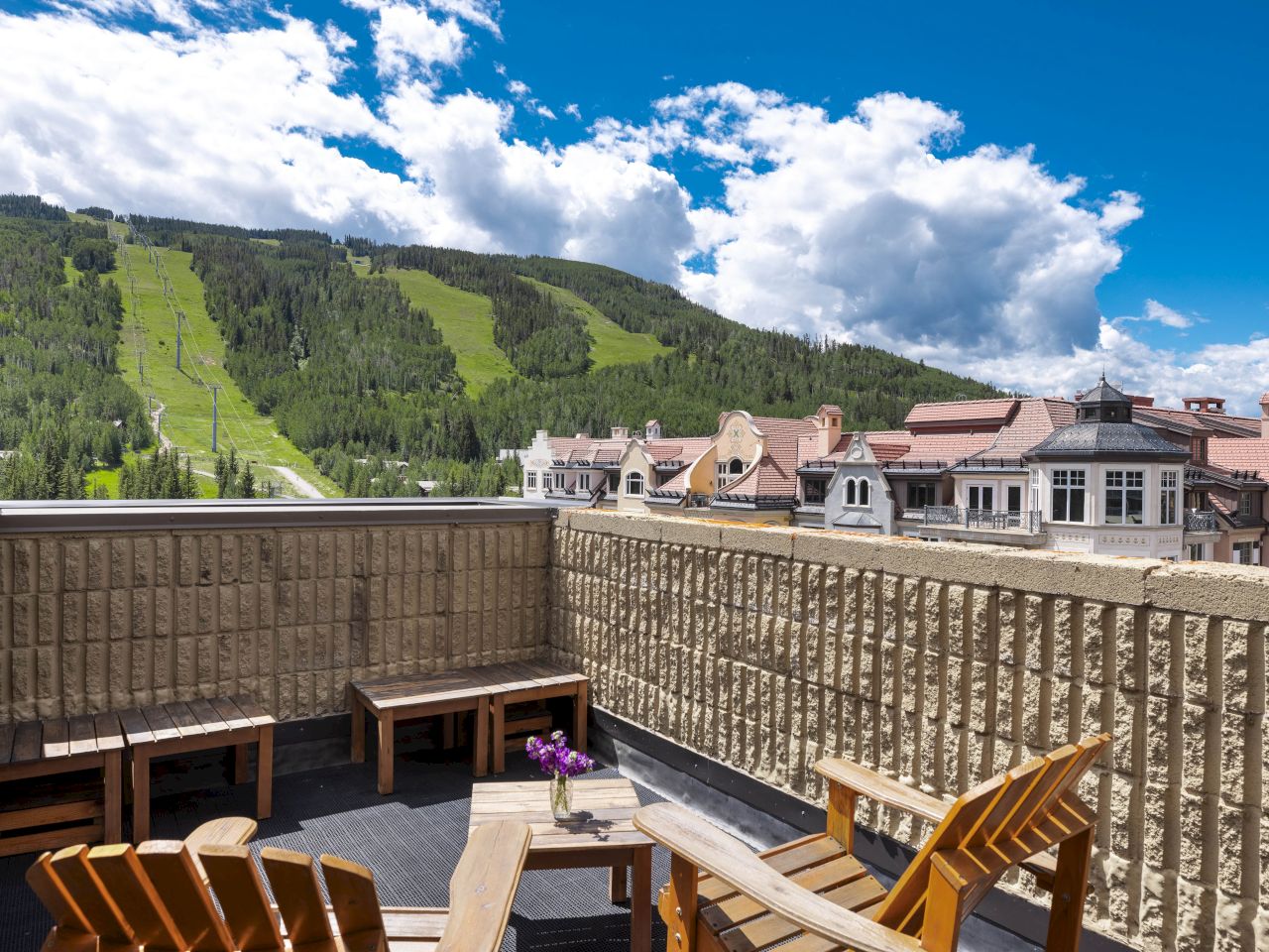 A scenic mountain view from a balcony with wooden chairs and tables, overlooking a quaint village under a blue sky with clouds.