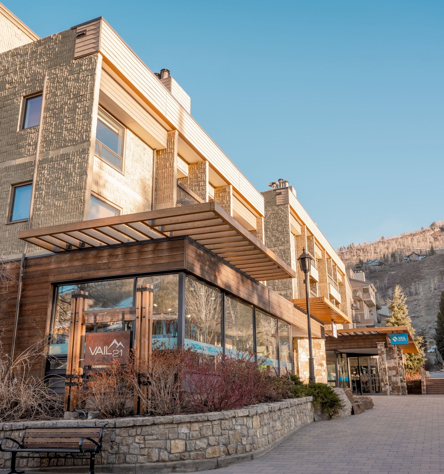 A modern building with large glass windows, brick and wood accents, surrounded by bare trees and a pathway, set against a clear sky.