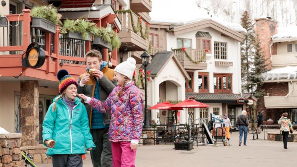 People in winter clothing enjoy snacks in a festive, snow-covered village street lined with buildings and winter decor.