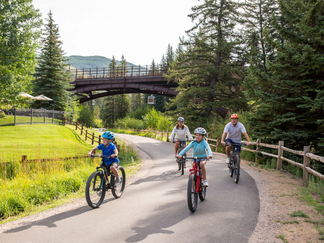 A group of people are biking on a paved path in a scenic area with trees, grass, and a bridge visible in the background.