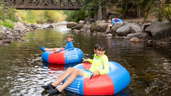 People are tubing on a river with a wooden bridge and trees in the background.