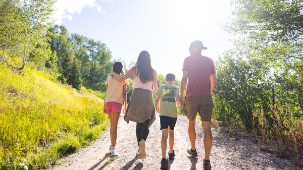 A family of four walking on a sunny path surrounded by greenery and trees, with the sun shining brightly in the background.