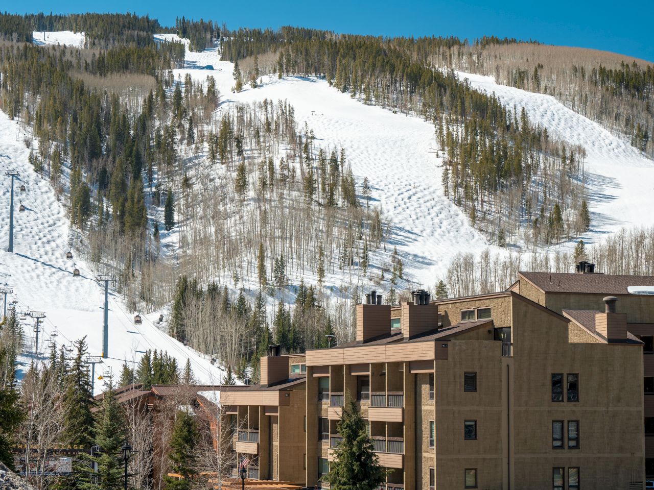 Snow-covered ski slopes with chairlifts on a mountain, surrounded by trees, and a building complex in the foreground.