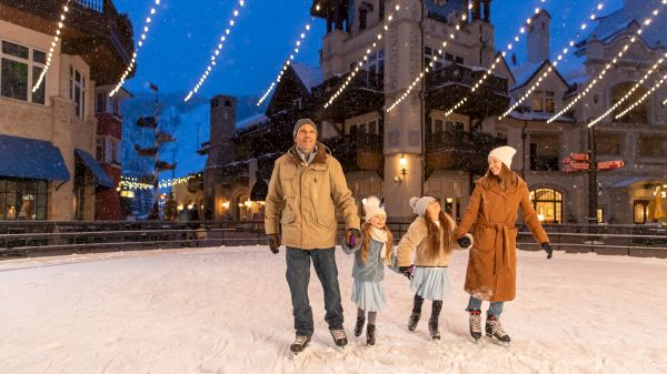 A family of four ice-skates outdoors under string lights in a festive town setting, with snow-covered buildings in the background.