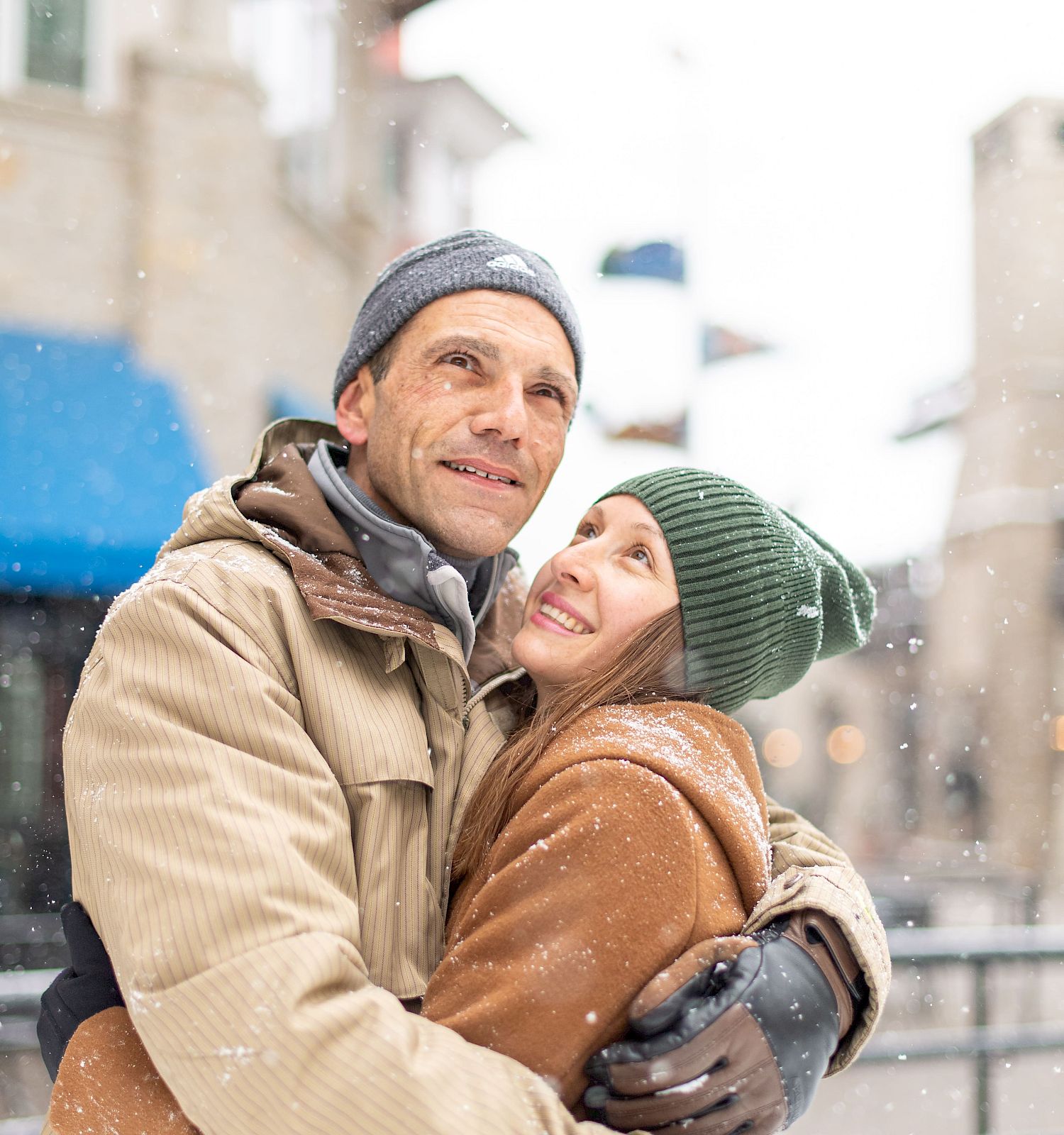 A couple wearing winter clothing hugs each other outside, surrounded by a snowy scene in an urban setting.