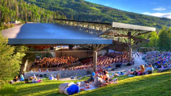 The image shows an outdoor amphitheater with people seated on the grass watching a performance, set against a backdrop of lush, green hills.