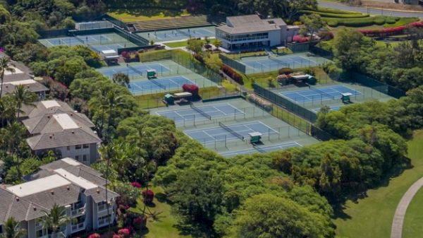 Aerial view of multiple tennis courts surrounded by greenery and buildings, featuring several courts arranged in rows with nearby facilities.