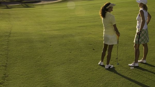 Two people stand on a golf course, holding clubs and wearing visors, amidst a sunlit landscape.