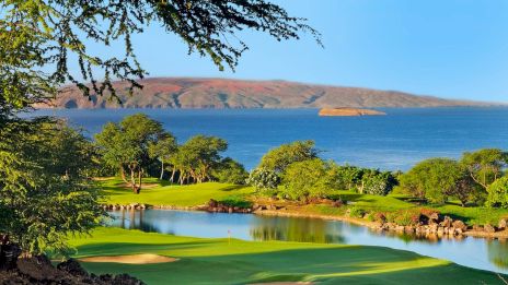 A scenic golf course with lush greenery, a sand trap, and a water hazard overlooks a calm ocean and distant landmass under a clear blue sky.