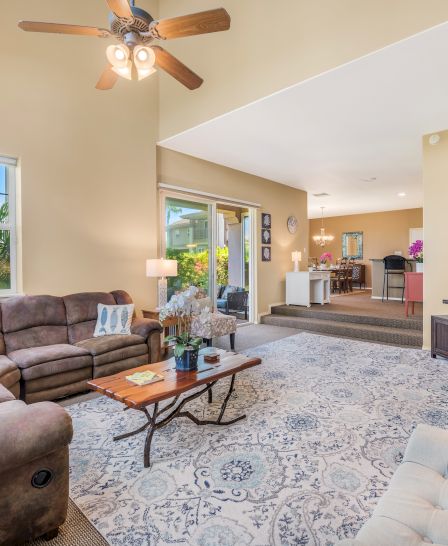 A cozy living room with plush sofas, a TV, a ceiling fan, large windows, and a patterned rug, opening into a dining area.