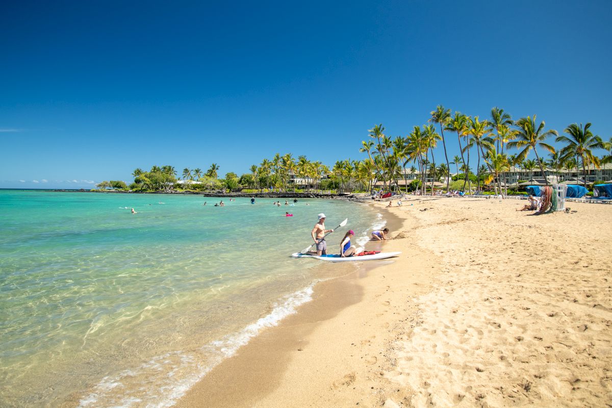 A sandy beach with clear turquoise water, palm trees, and people enjoying the sun and water activities under a clear blue sky.
