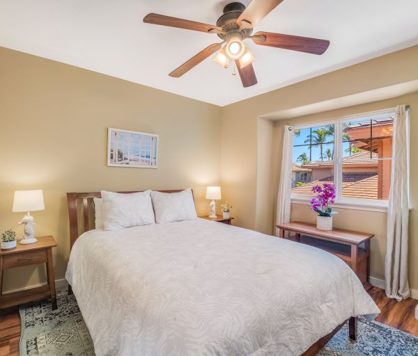 A cozy bedroom with a bed, nightstands, lamps, a ceiling fan, and a window with a potted plant on a table.