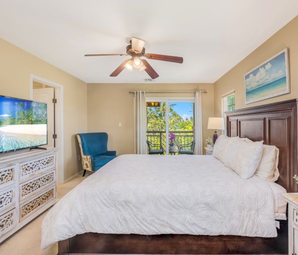 A cozy bedroom features a large bed, TV, chair, ceiling fan, and balcony view, with beach-themed decor and natural light filling the space.
