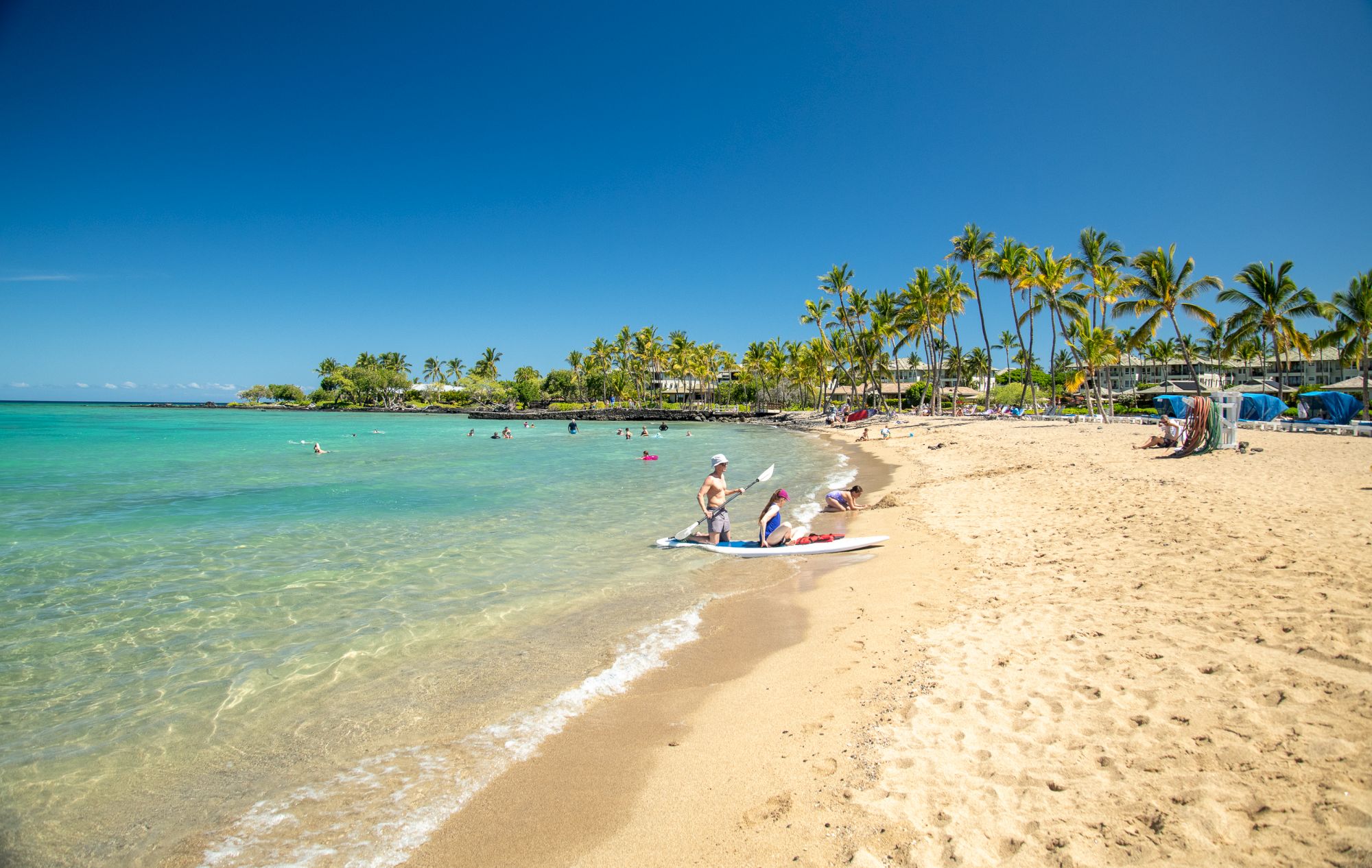 A tropical beach with clear turquoise water, golden sand, palm trees, and people enjoying a sunny day near a small boat by the shore.