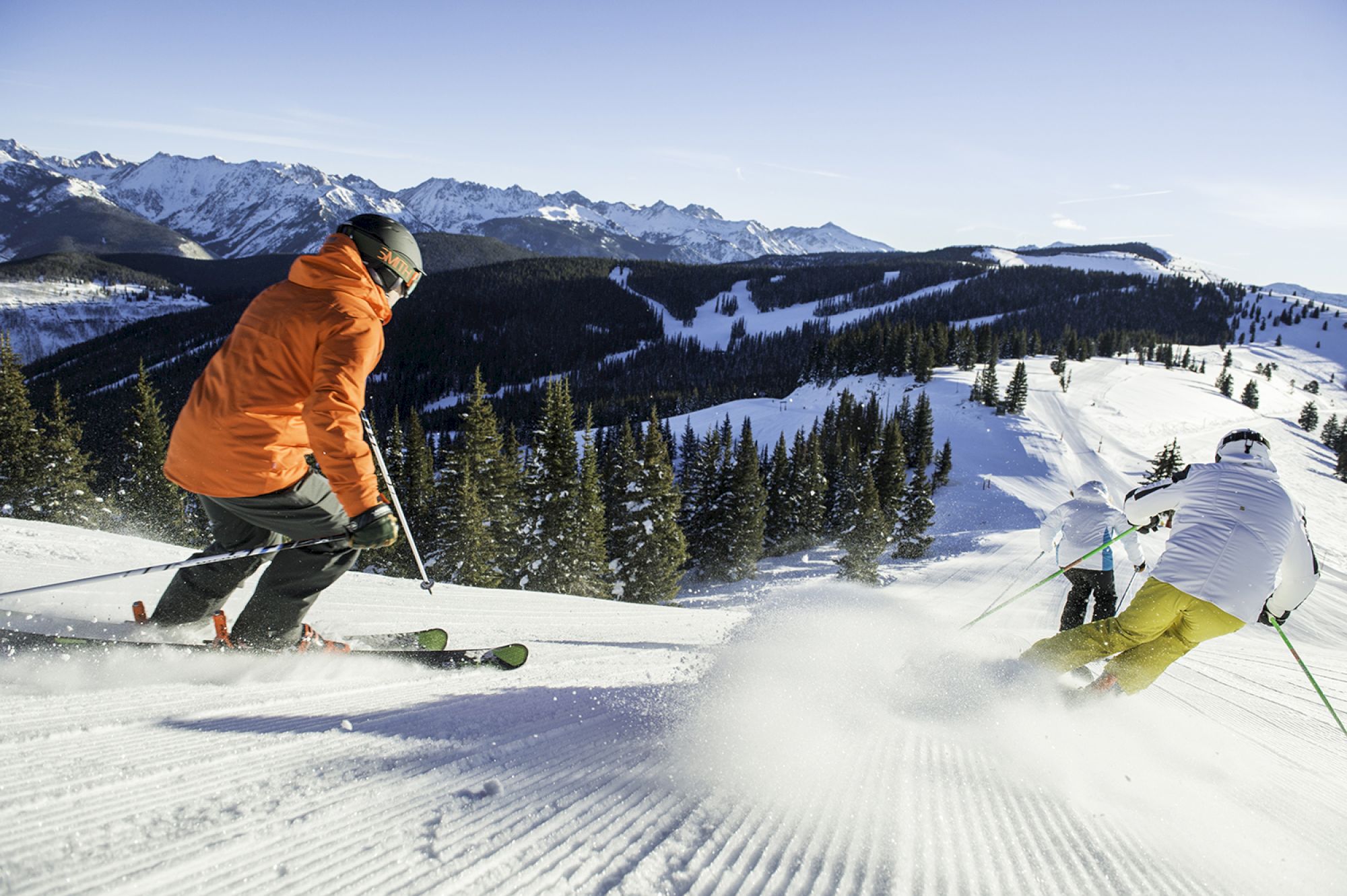 The image shows skiers enjoying a snowy mountain slope under a clear blue sky, surrounded by trees and impressive peaks.