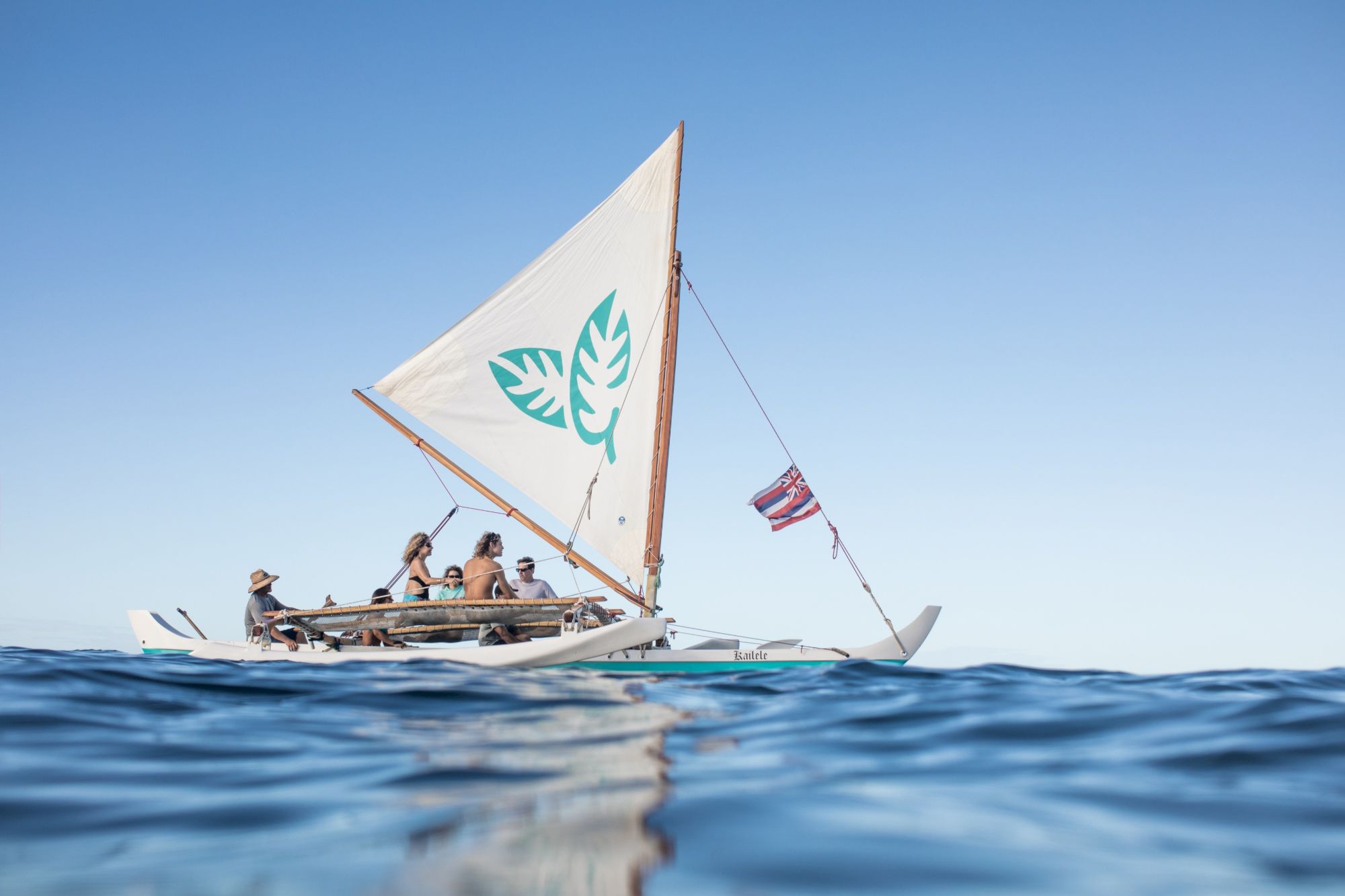 A canoe with a large sail and several people is sailing on calm water under a clear blue sky, creating a serene outdoor atmosphere.