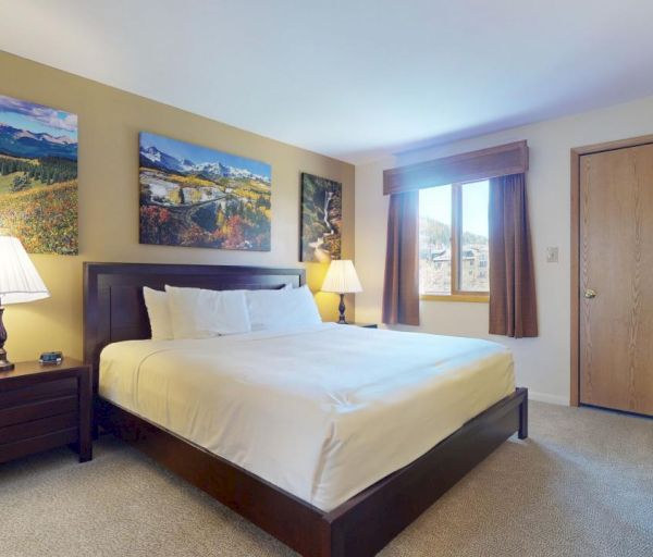 The image shows a cozy bedroom featuring a large bed, lamp, artwork, and a view through a window, all in warm tones.