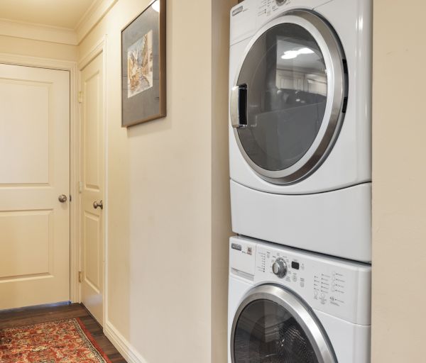 The image shows a hallway with a stacked washer and dryer and a door leading to another room, along with a framed picture.