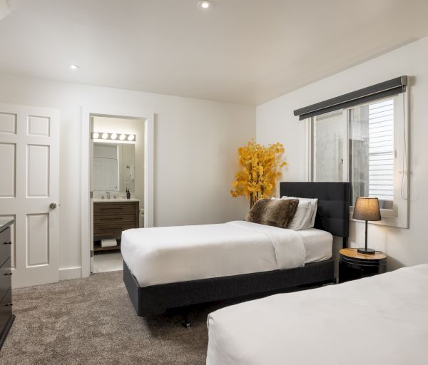 The image shows a cozy bedroom with two beds, a dresser, a lamp, and a bathroom doorway, featuring a touch of yellow decor.
