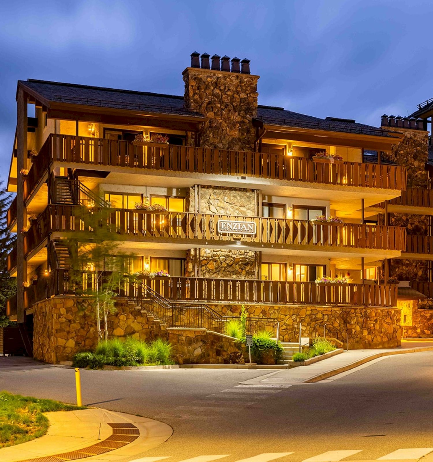 The image shows a rustic-style multi-story building at dusk with wooden balconies and lit pathways.