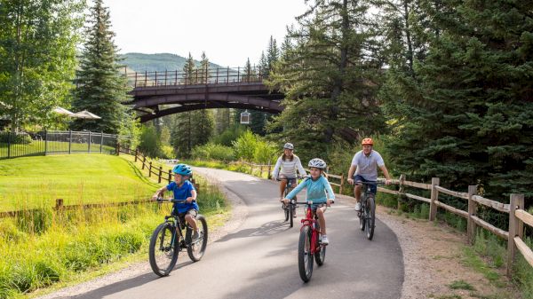 A family is biking on a paved path surrounded by greenery, trees, and a bridge in the background.