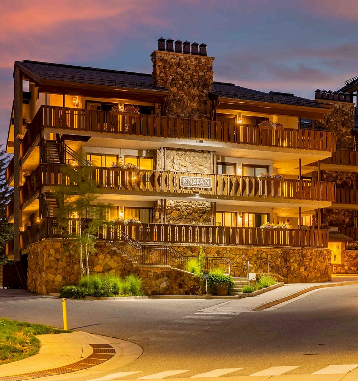 The image shows a cozy, well-lit lodge with a stone facade, surrounded by trees and a gently winding path. It exudes a warm ambiance.