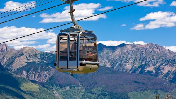 The image features a gondola cable car traveling high above lush green mountains under a bright blue sky with scattered clouds.