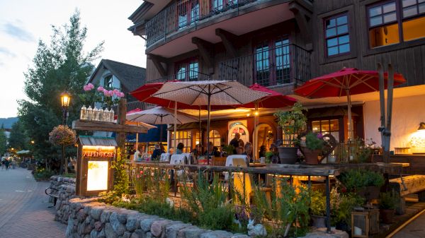 A cozy restaurant with outdoor seating, umbrellas, flowers, and warm lighting, creating an inviting atmosphere for diners.