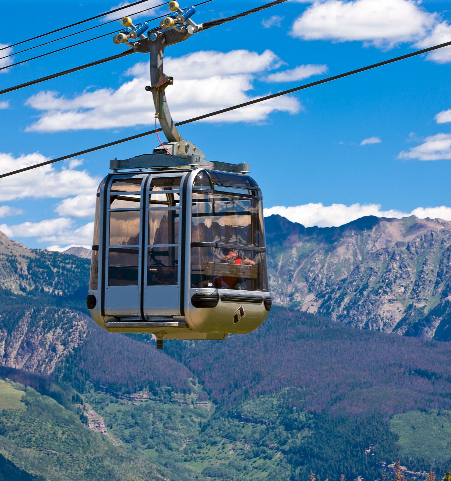 A gondola lift travels along a cable with scenic mountains and a bright blue sky in the background.