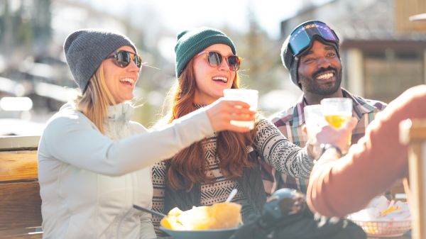 A group of friends cheers with drinks outdoors, enjoying a sunny day after skiing, with food and smiles all around.