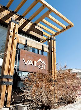 The image shows the entrance to "Vail 21" with wooden accents, nearby "Vail Sports," and two people walking with ski gear in sunny weather.