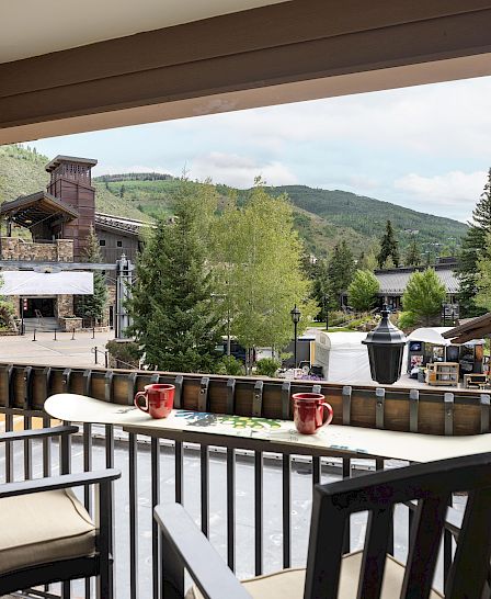 A balcony with two chairs and mugs overlooks a scenic mountain view with trees and a rustic building in the background.