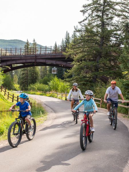 A family of four is cycling on a scenic paved trail, surrounded by trees and near a wooden bridge, on a sunny day.