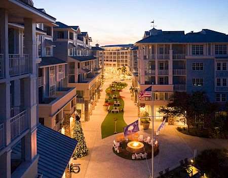 The image shows a beautifully lit courtyard between modern buildings, featuring a fire pit, greenery, and festive decorations.