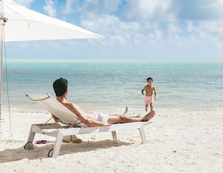 A man lounging on a beach chair under a white umbrella watching a child playing near the turquoise sea, with clear blue sky above.